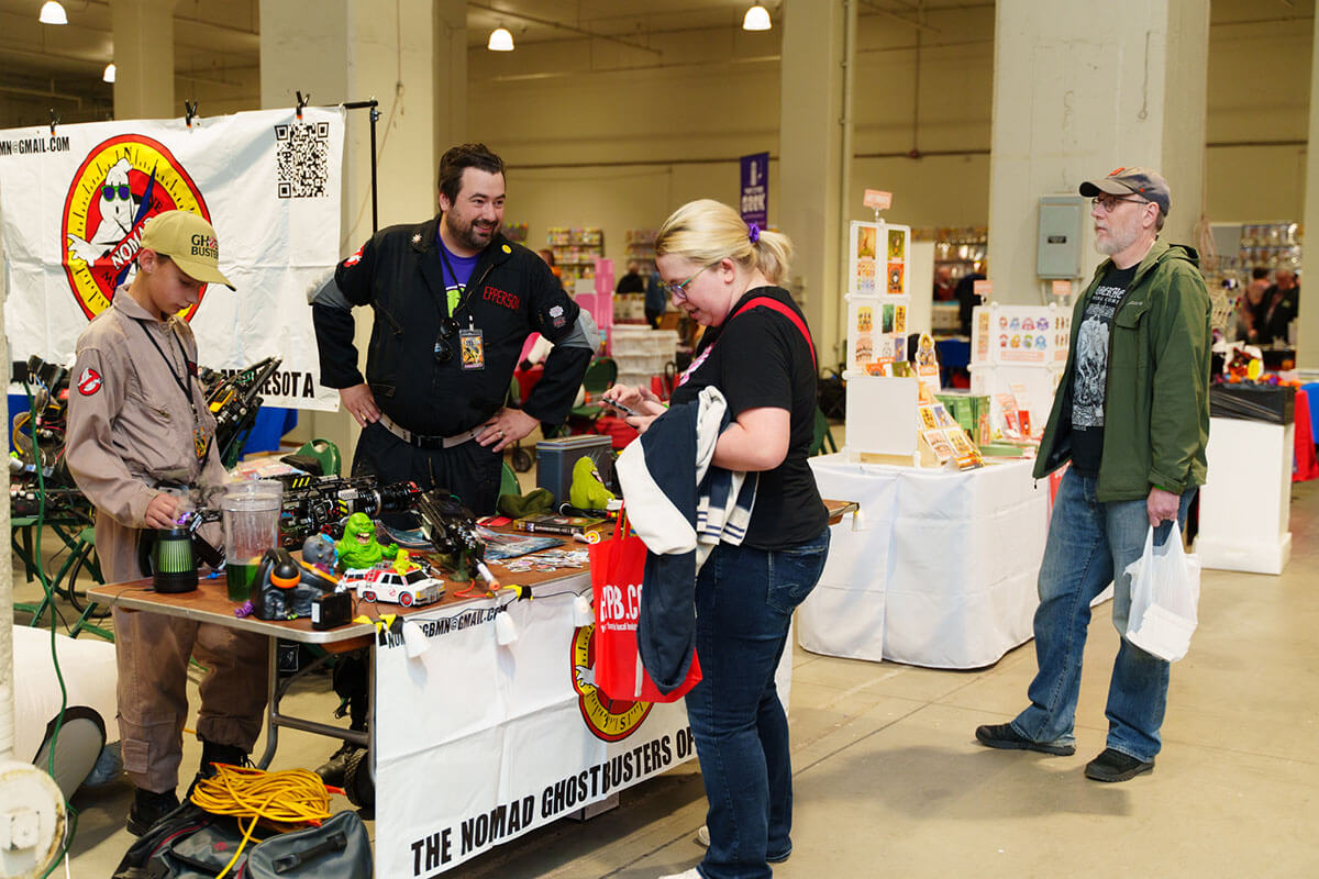 The NOMAD Ghostbusters at MNCBA FallCon 2024. Photo by Buzz Meade. two people holding yellow plastic bags full of comic books and handing them out to attendees at admissions