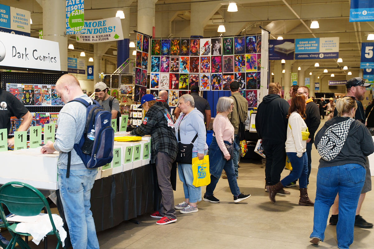 Attendees browsing a MNCBA FallCon 2024 creator and vendor booths. Photo by Buzz Meade. a small crowd of attendees searching comic book shortboxes in the foreground with a tall creator backdrop of prints in the background