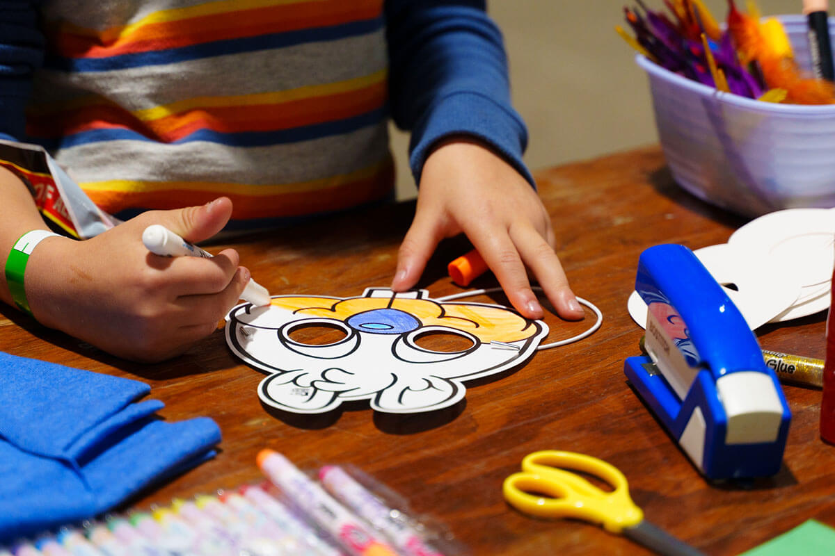 Child making a Shortbox the Squirrel mask at MNCBA FallCon 2024. Photo by Buzz Meade. photo of a child colouring a paper mask with markers on a brown tabletop